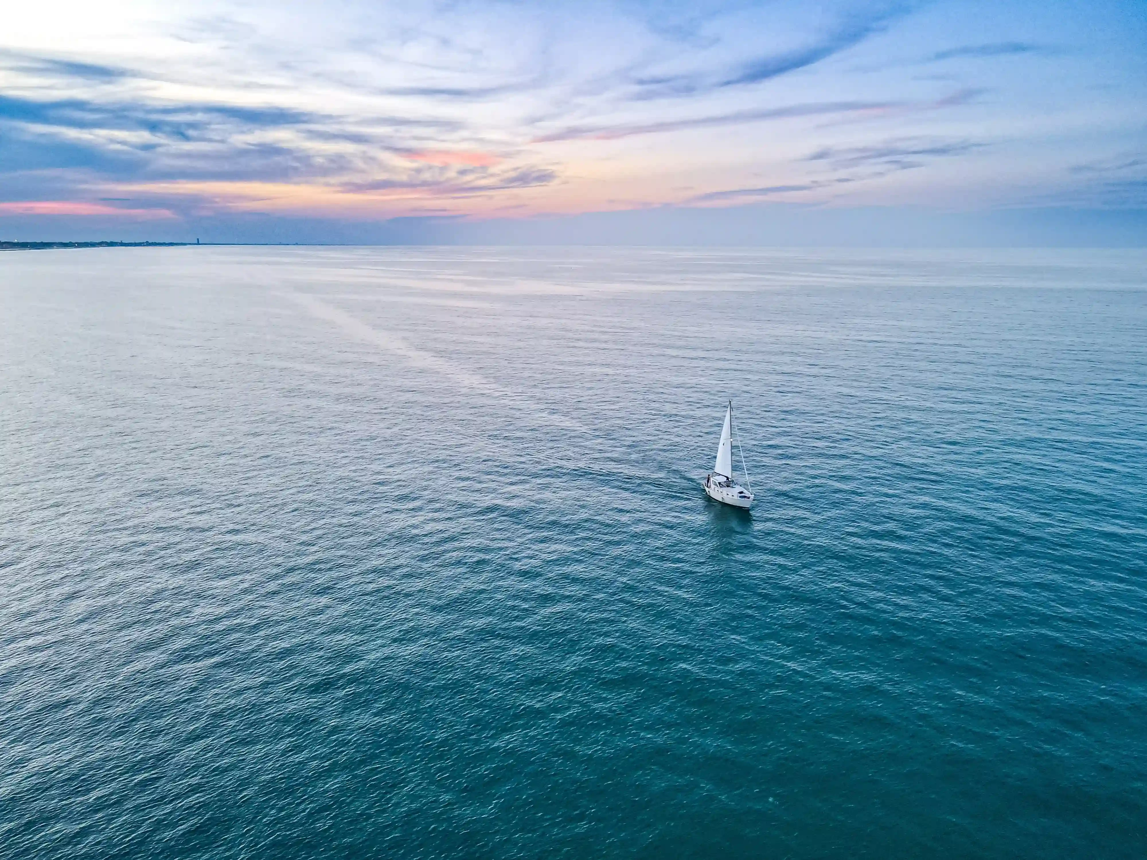 Vue aérienne d'un voilier naviguant sur une mer calme au coucher du soleil, avec un vaste horizon et un ciel aux nuances bleues et rosées, idéale pour illustrer une location de voilier ou une croisière en mer.