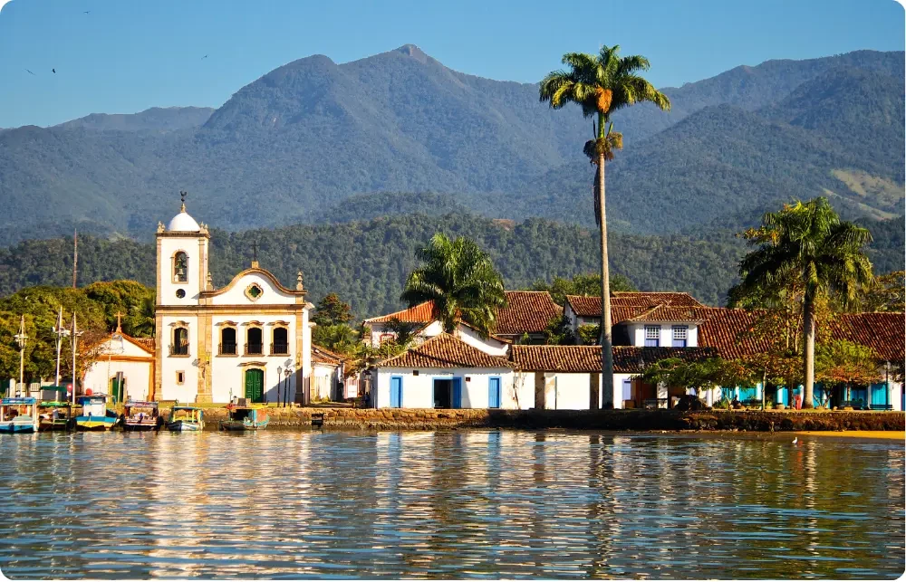 Vue depuis la baie sur le centre historique de Paraty au Br&eacute;sil : &eacute;glise baroque coloniale, maisons color&eacute;es aux volets bleus, palmiers et montagne de la Serra da Bocaina en arri&egrave;re-plan