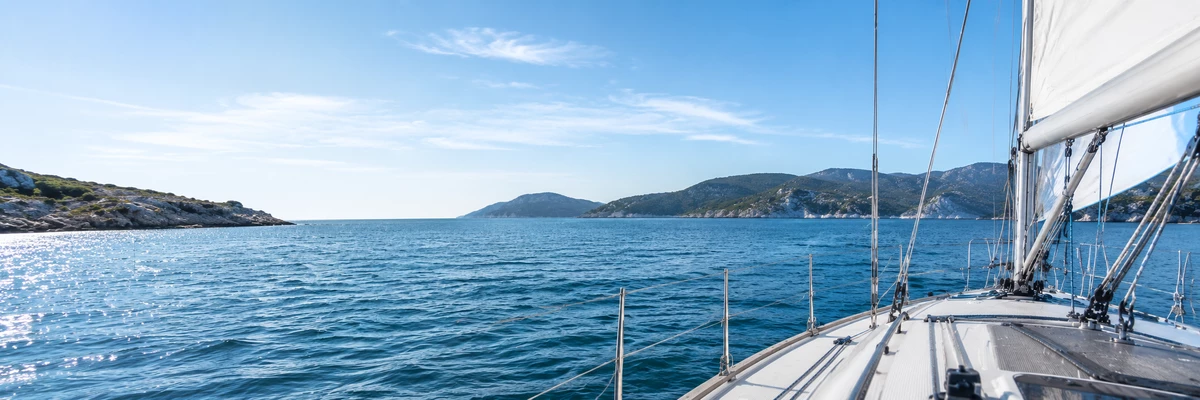 Vista desde la cubierta de un velero que navega entre las islas del Mediterráneo, con sus aguas turquesas bajo un cielo azul y luminoso.