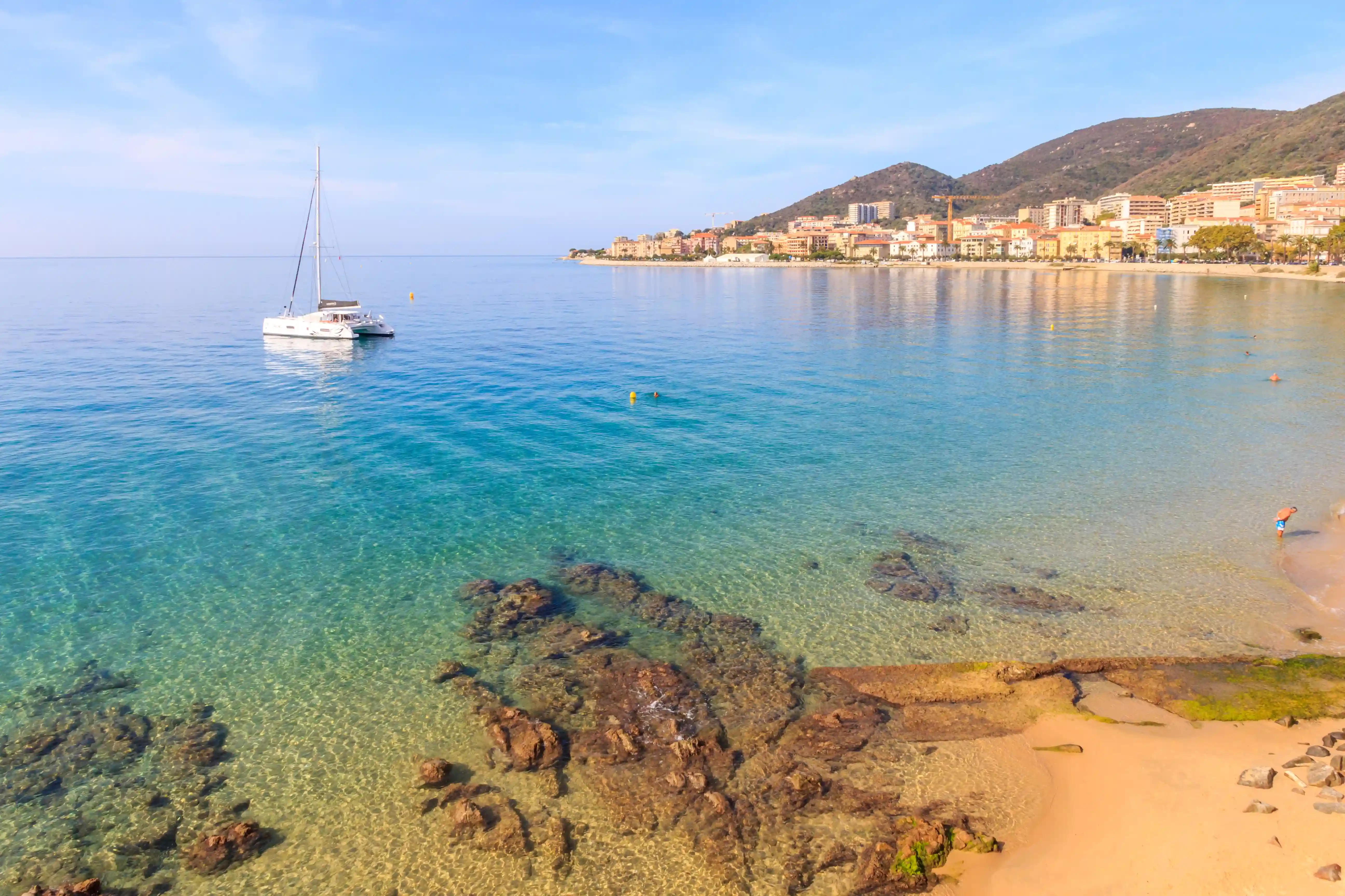 Catamaran au mouillage dans une baie aux eaux turquoise lors d'une croisi&egrave;re d'une semaine. Catamaran naviguant pr&egrave;s du littoral dans une baie aux eaux transparentes, avec une ville c&ocirc;ti&egrave;re et des montagnes en arri&egrave;re-plan.