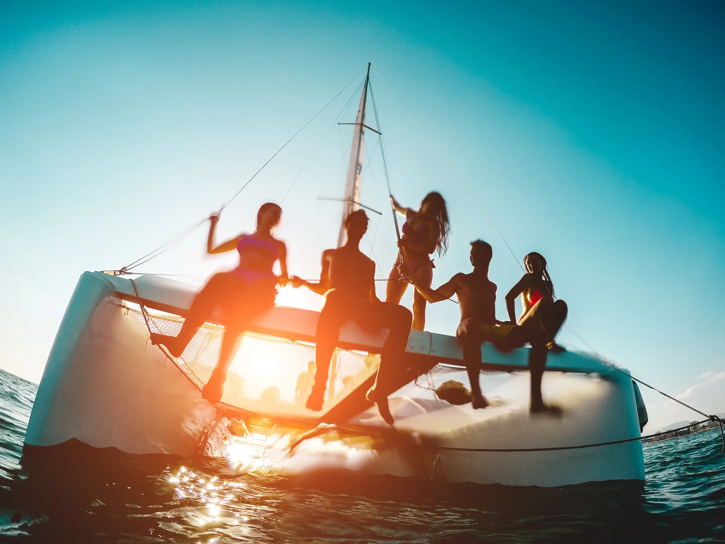 Groupe d'amis sur le trampoline d'un catamaran privatis&eacute; pour une croisi&egrave;re d'une semaine. Groupe d&rsquo;amis profitant d&rsquo;un moment de d&eacute;tente &agrave; bord d&rsquo;un catamaran au coucher du soleil, les pieds au-dessus de l&rsquo;eau dans une ambiance festive et conviviale.