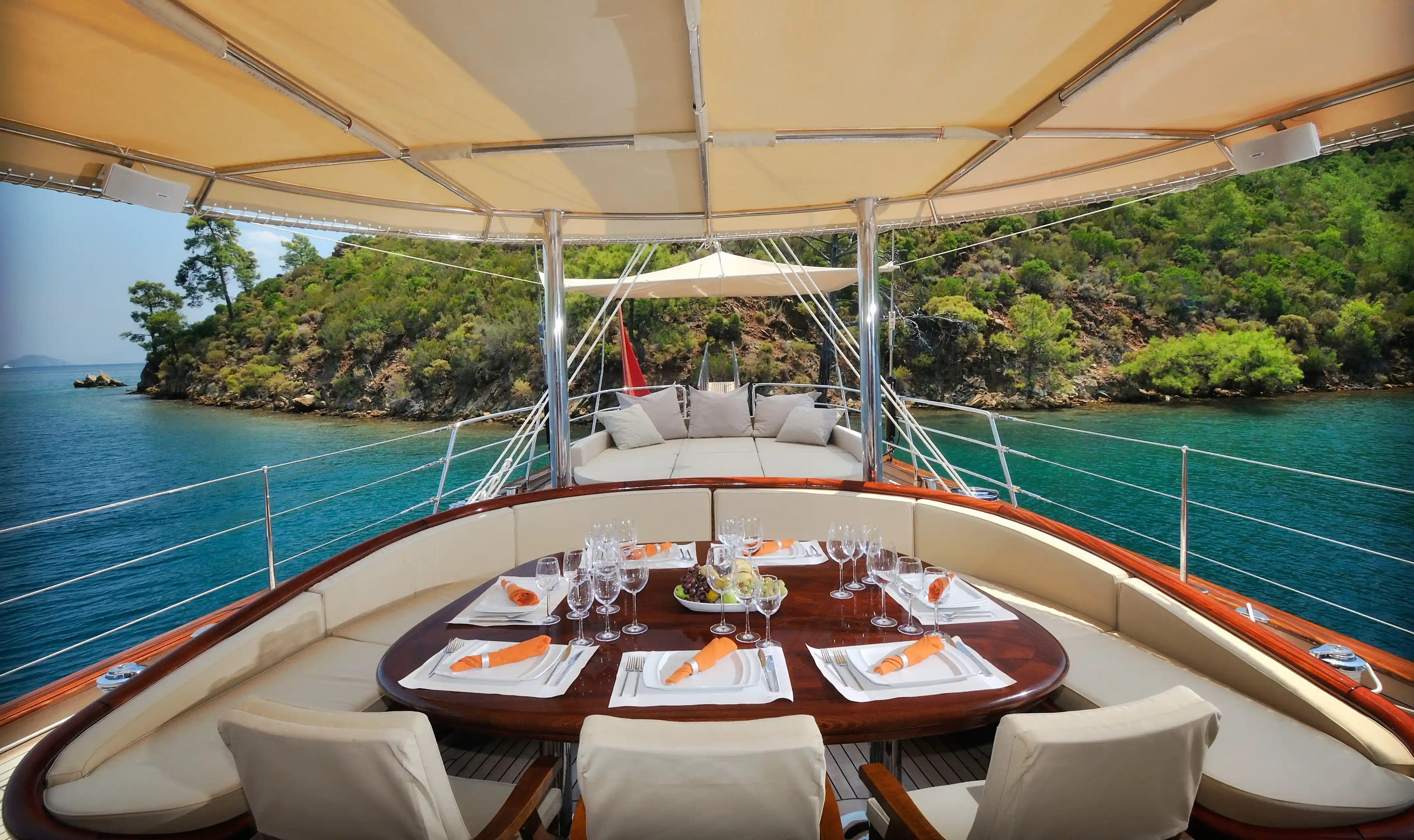 D&icirc;ner convivial sur le pont d'un voilier au coucher du soleil pendant une croisi&egrave;re. Table dress&eacute;e sur le pont d&rsquo;un yacht avec vue sur une crique aux eaux turquoise, offrant une exp&eacute;rience de repas en plein air dans un cadre intimiste et haut de gamme.