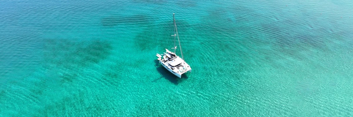 Vue aérienne d’un catamaran blanc au mouillage dans une eau turquoise cristalline. Le bateau est entouré de nuances de bleu et de vert, révélant les fonds marins peu profonds d’un lagon tropical sous un ciel dégagé et une lumière de pleine journée.