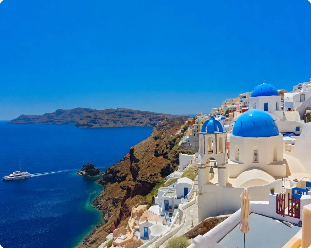 Village d'Oia à Santorin dans les Cyclades, maisons blanches et églises aux dômes bleus surplombant la caldeira, bateau naviguant sur la mer Égée bleu profond