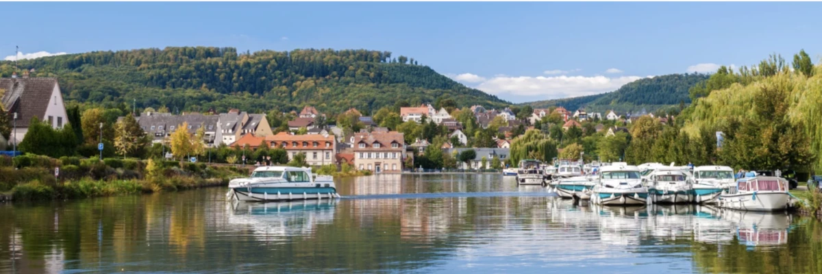 Hausboote auf dem Rhein-Marne-Kanal vor einem elsässischen Dorf mit bewaldeten Hügeln
