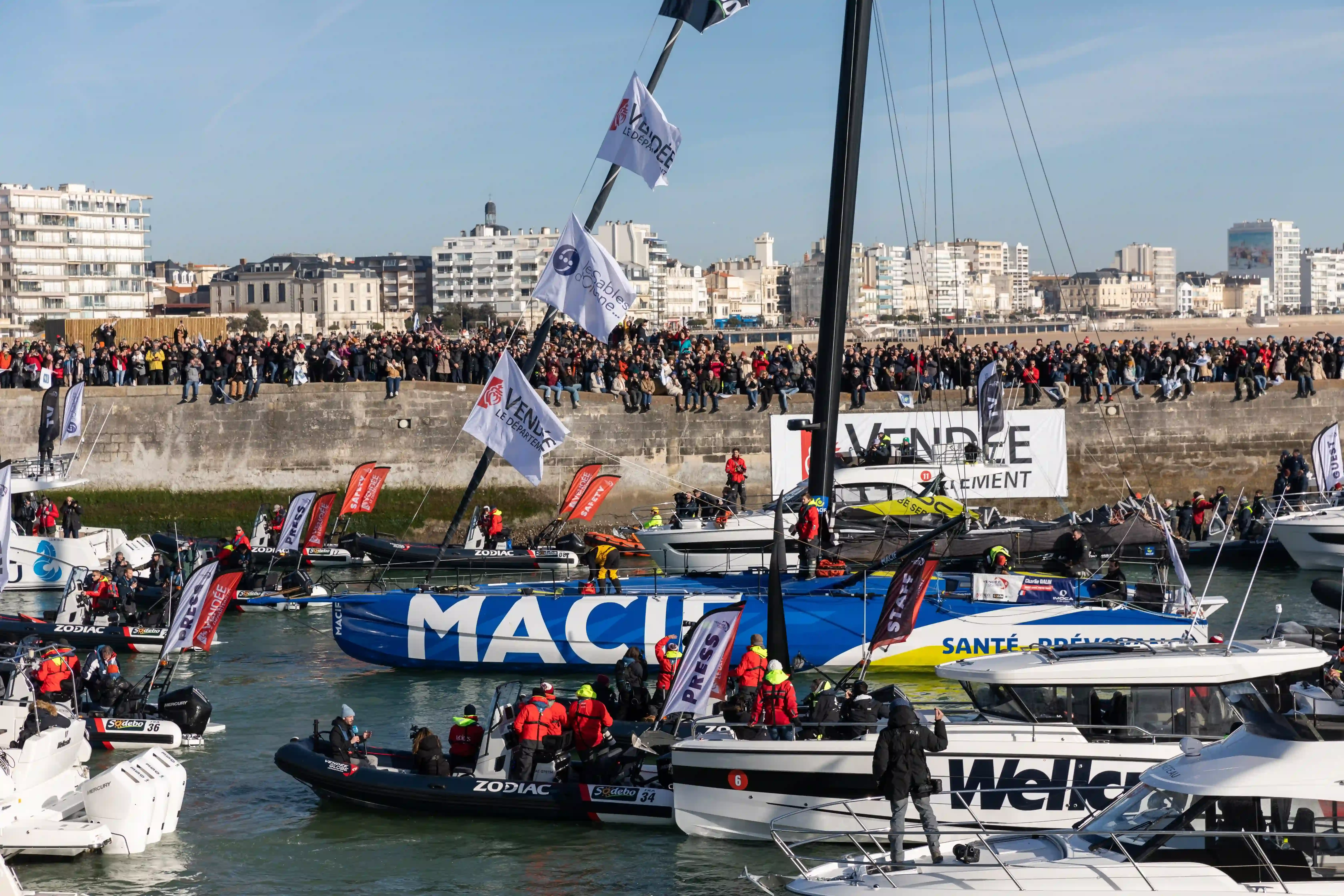 le voilier IMOCA bleu et jaune Macif Santé Prévoyance de Charlie Dalin remonte le chenal des Sables-d'Olonne, escorté par une nuée de bateaux suiveurs, sous les acclamations d'une foule immense massée sur les quais