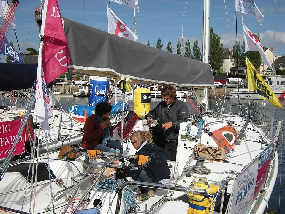 Charlie Dalin, en veste sombre, debout dans le cockpit de son voilier Figaro à quai lors de la Solitaire du Figaro 2012 à Paimpol, en discussion avec deux personnes au milieu des cordages, entouré de pavillons de course