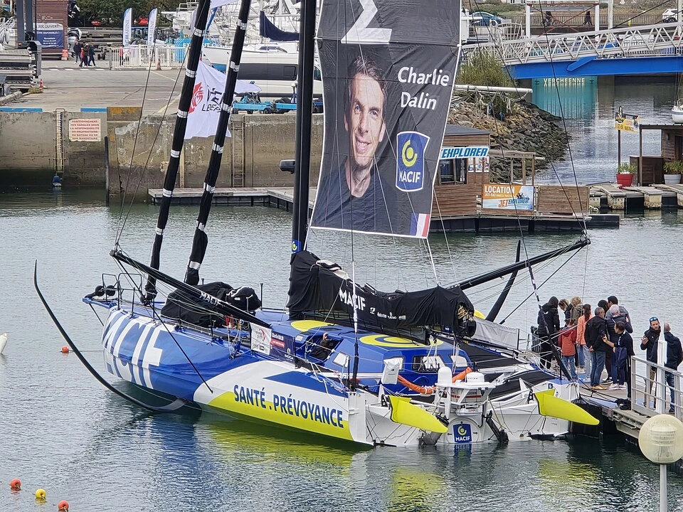 l'IMOCA Macif Santé Prévoyance de Charlie Dalin amarré à un ponton, une grande voile dressée portant le portrait du skipper et le logo MACIF, des visiteurs observant le bateau depuis le quai