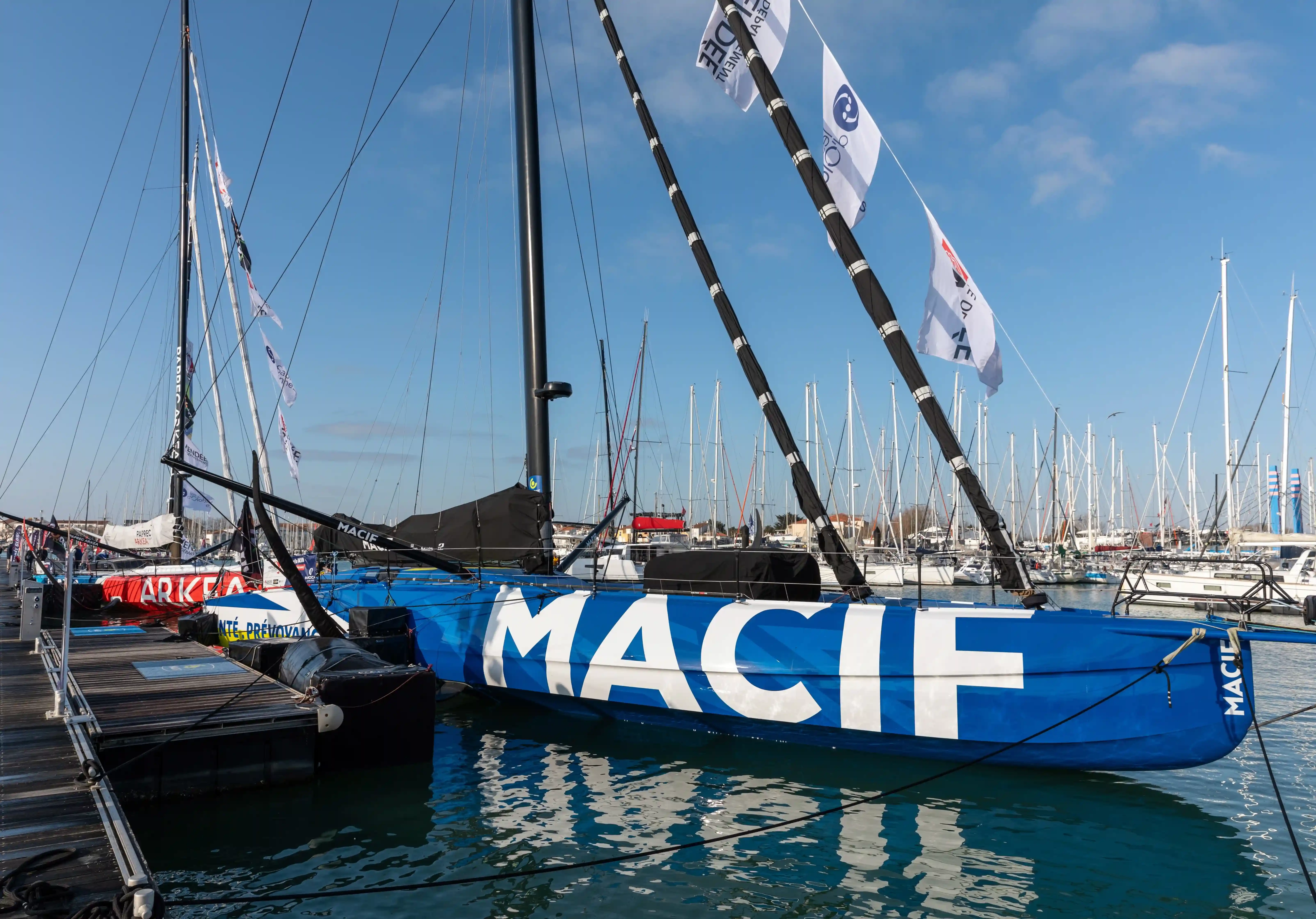 l'IMOCA 60 bleu et blanc Macif Santé Prévoyance de Charlie Dalin amarré à un ponton dans un port, voiles affalées et grand mât dressé, au milieu d'une forêt de mâts de voiliers sous un ciel bleu