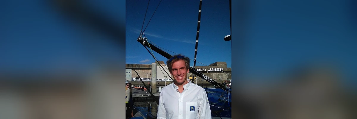 portrait souriant de Charlie Dalin en chemise blanche au logo MACIF, des lunettes de soleil relevées sur le front, devant l'étrave de son IMOCA sur le ponton de Lorient La Base, sur un fond bleu flouté en bannière