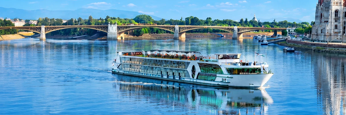 Bateau de croisière fluviale naviguant sur le Danube à Budapest, avec le Parlement hongrois et un pont historique en arrière-plan, sous un ciel bleu lumineux se reflétant dans les eaux calmes du fleuve.