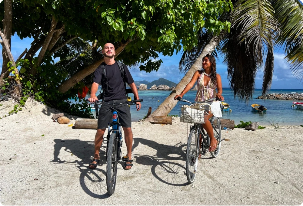 Deux voyageurs à vélo sur une plage des Seychelles, entre palmiers, mer turquoise et bateaux au mouillage, dans un décor tropical baigné de soleil.