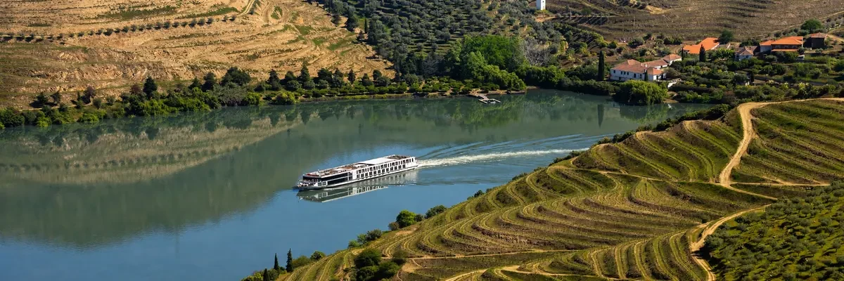 Luftaufnahme eines modernen Flusskreuzfahrtschiffs auf dem Douro, das durch ruhiges Wasser zwischen terrassenförmig angelegten Weinbergen und ländlicher Landschaft unter klarem Himmel fährt.