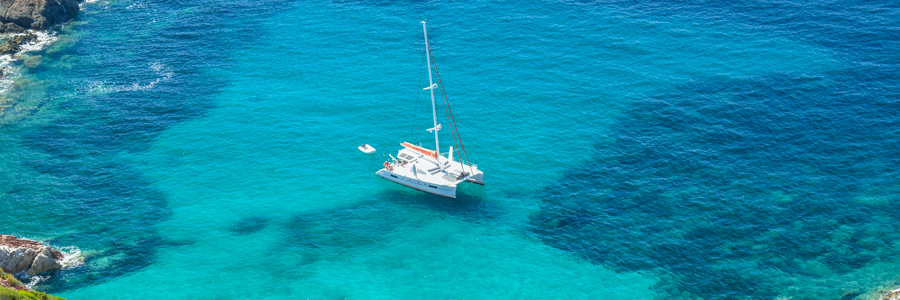 Catamarano all'ancora in acque turchesi e cristalline al largo di una costa rocciosa del Mediterraneo, visto dall'alto durante una crociera estiva.